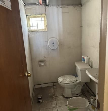 Baño pequeño y deteriorado en propiedad comercial, con techo incompleto, ventilación natural y accesorios básicos para uso ocasional. Ideal para remodelar o invertir en bienes raíces comerciales.