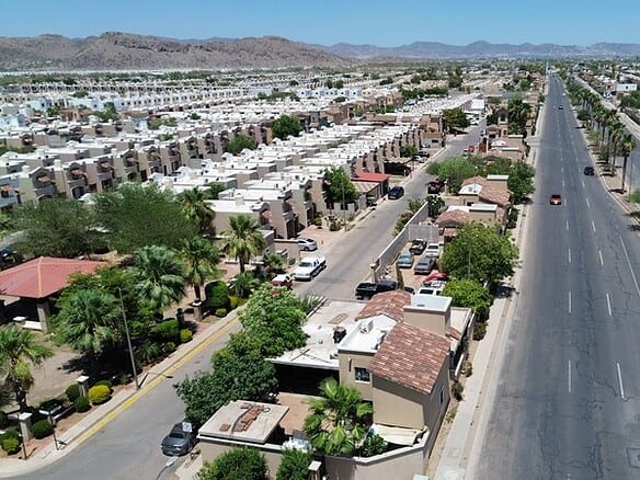 Vista aérea de una urbanización moderna con viviendas residenciales y áreas comerciales en un entorno urbano, mostrando calles amplias, áreas verdes y un paisaje montañoso al fondo.
