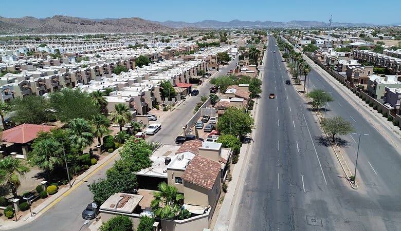 Vista aérea de una urbanización moderna con viviendas residenciales y áreas comerciales en un entorno urbano, mostrando calles amplias, áreas verdes y un paisaje montañoso al fondo.